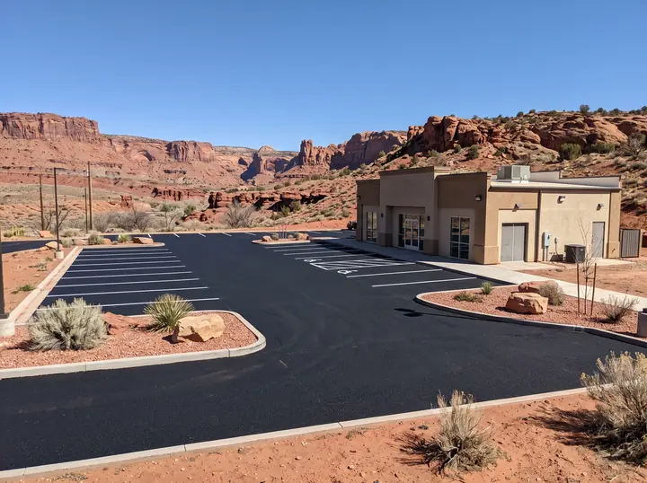 Newly paved asphalt parking lot with fresh white stall lines at a stucco commercial building below red sandstone cliffs